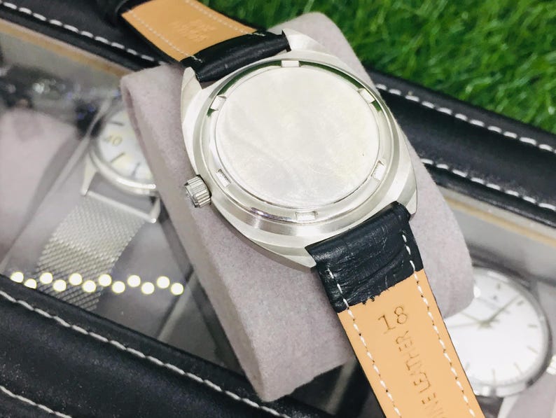 Op de afbeelding: Close-up van een zilverkleurig polshorloge met een zwarte leren band. Het horloge staat tegen een grijze achtergrond, met andere horloges zichtbaar op de achtergrond. De band is gemarkeerd met "GENUINE LEATHER" en het nummer "18".