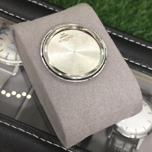 May include: A silver watch case back with the text "100% WATERPROOF LE COULTRE CO SWISS" is displayed on a gray cushion inside a black watch case. Other watches are visible in the background.
