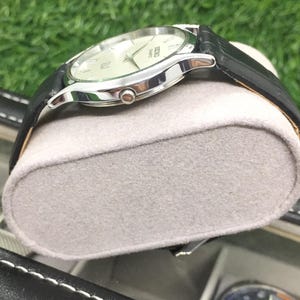 May include: A silver-toned wristwatch with a black leather band. The watch has a light-colored face with silver hands and markers. The watch is resting on a gray, oval-shaped cushion.