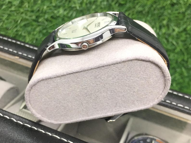 May include: A silver-toned wristwatch with a black leather band. The watch has a light-colored face with silver hands and markers. The watch is resting on a gray, oval-shaped cushion.