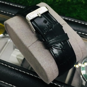 K&ouml;nnte beinhalten: Ein schwarzes Leder-Uhrenarmband mit einer silbernen Schnalle, das den Markennamen "SEIKO" tr&auml;gt. Das Armband wird auf einem grauen Kissen in einem schwarzen Uhrenetui pr&auml;sentiert. Das Armband hat eine strukturierte Oberfl&auml;che.