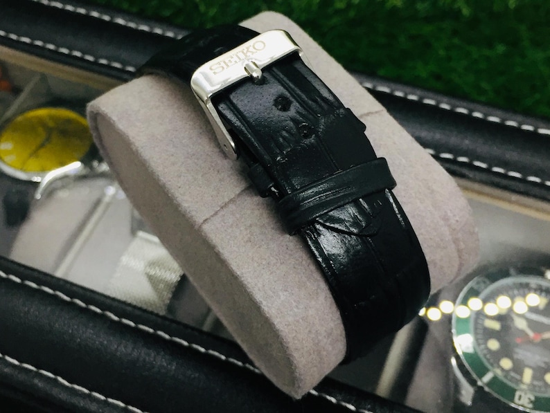 K&ouml;nnte beinhalten: Ein schwarzes Leder-Uhrenarmband mit einer silbernen Schnalle, das den Markennamen "SEIKO" tr&auml;gt. Das Armband wird auf einem grauen Kissen in einem schwarzen Uhrenetui pr&auml;sentiert. Das Armband hat eine strukturierte Oberfl&auml;che.
