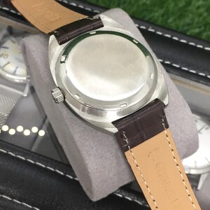 May include: Close-up of a silver-toned watch with a brown leather strap. The watch is displayed in a black case with other watches visible in the background. The watch has a silver-colored dial and a stainless steel back.