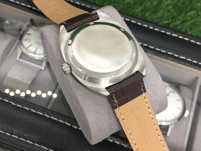 May include: Close-up of a silver-toned watch with a brown leather strap. The watch is displayed in a black case with other watches visible in the background. The watch has a silver-colored dial and a stainless steel back.