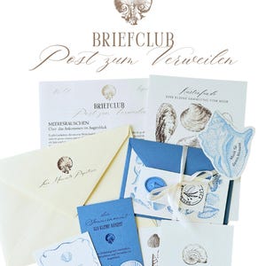 Puede incluir: Un conjunto de papelería con sobres crema y azules, tarjetas con ilustraciones de conchas marinas y texto en alemán que incluye BRIEFCLUB, Post zum Verweilen y Meeresrauschen. Un sello de cera azul y una cinta blanca decoran el sobre azul.
