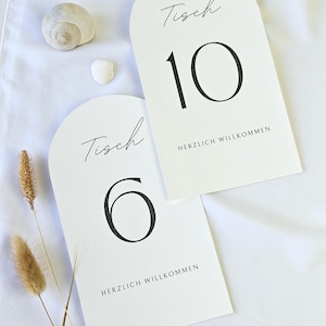 May include: Two white arched table number cards with black text. One card displays the number 10, the word "Tisch", and "HERZLICH WILLKOMMEN". The other card shows the number 6, the word "Tisch", and "HERZLICH WILLKOMMEN".