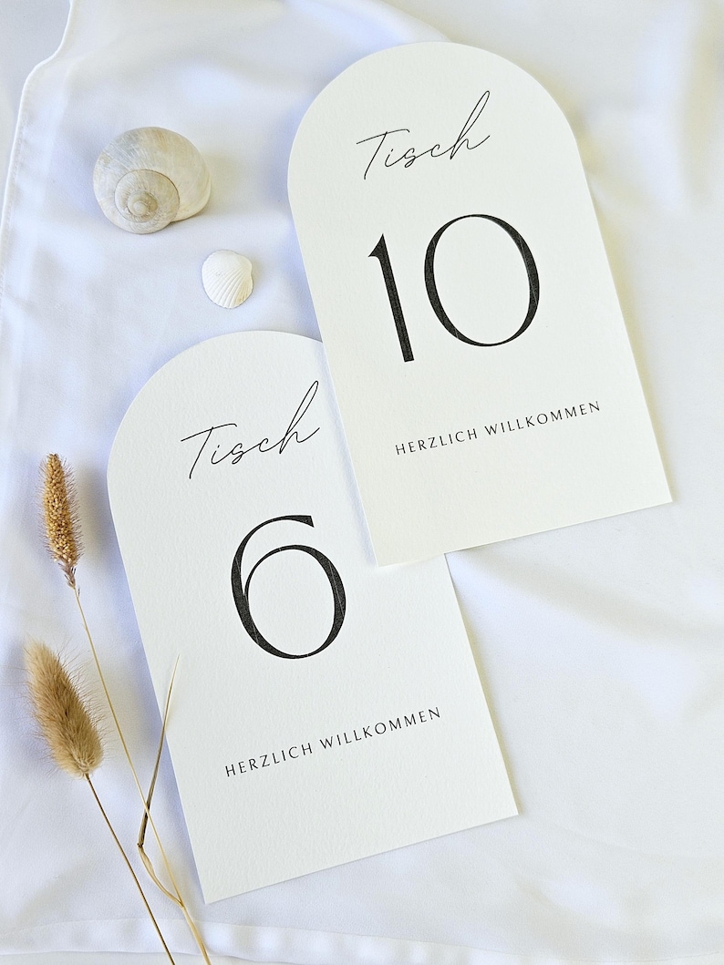 May include: Two white arched table number cards with black text. One card displays the number 10, the word "Tisch", and "HERZLICH WILLKOMMEN". The other card shows the number 6, the word "Tisch", and "HERZLICH WILLKOMMEN".