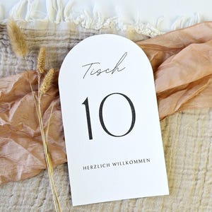 May include: White arched table number card with the number 10 in black. The word "Tisch" is written in script above the number, and "HERZLICH WILLKOMMEN" is printed below. The card is on a textured surface.