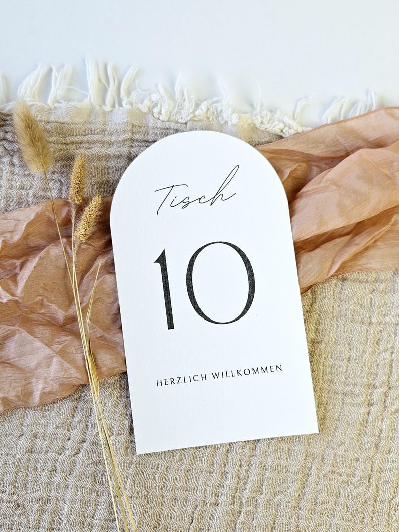 May include: White arched table number card with the number 10 in black. The word "Tisch" is written in script above the number, and "HERZLICH WILLKOMMEN" is printed below. The card is on a textured surface.