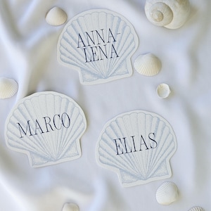 May include: Three seashell-shaped place cards with names printed in navy blue: ANNA-LENA, MARCO, and ELIAS. Several small seashells and a large spiral shell are scattered around the cards on a white fabric background. The place cards have a light blue seashell design.