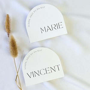 Könnte beinhalten: Zwei weiße, bogenförmige Tischkarten mit schwarzem Text. Auf einer Karte steht "MARIE" und auf der anderen "VINCENT". Beide Karten tragen den Satz "SCHON, DASS DU DA BIST". Links ist ein getrockneter Pflanzenstiel.