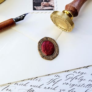 May include: An overhead shot of stationery items. A gold wax seal with a wooden handle sits next to a red wax seal. A pen and a stamp are also visible. The stamp reads "50 Loca Papeterie".