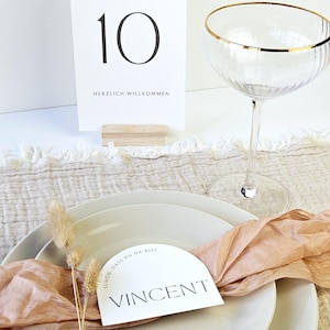 May include: A table setting with a white place card that reads "Vincent" and a table number card that reads "Tisch 10". The setting includes a champagne glass with a gold rim, white plates, and a peach-colored ribbon.