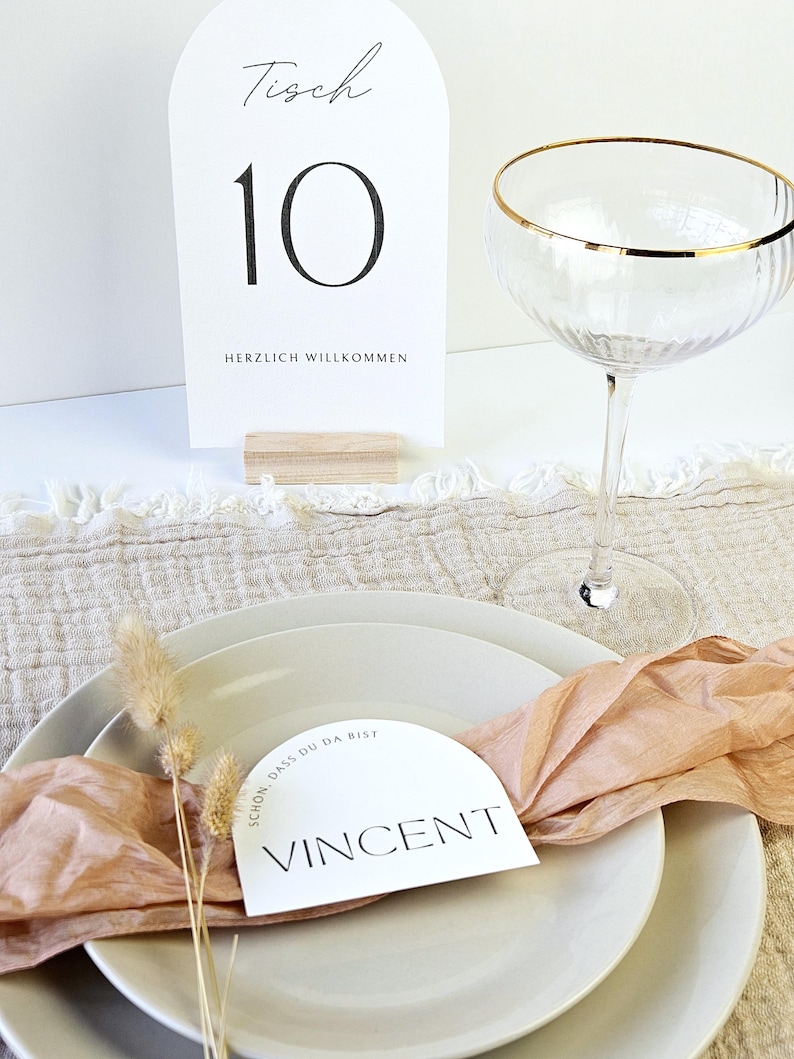 May include: A table setting with a white place card that reads "Vincent" and a table number card that reads "Tisch 10". The setting includes a champagne glass with a gold rim, white plates, and a peach-colored ribbon.
