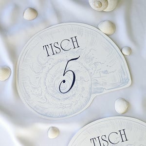 Könnte beinhalten: Weiße tischkarten in Muschelform mit dem Wort "TISCH" und einer Zahl in Marineblau. Die Karten sind von kleinen Muscheln auf weißem Stoffhintergrund umgeben. Ideal für eine Veranstaltung zum Thema Strand.