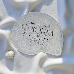 May include: A white, seashell-shaped invitation with the text "Save the Date" and the names "CAROLINA & RAFAEL" along with the date "23.07.2027". A small seashell rests beside the invitation on a white, textured surface.