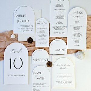 May include: A collection of white wedding stationery with black text, including invitations, menus, and table cards. The cards feature arched tops and elegant fonts. Decorative elements include wax seals and a seashell.