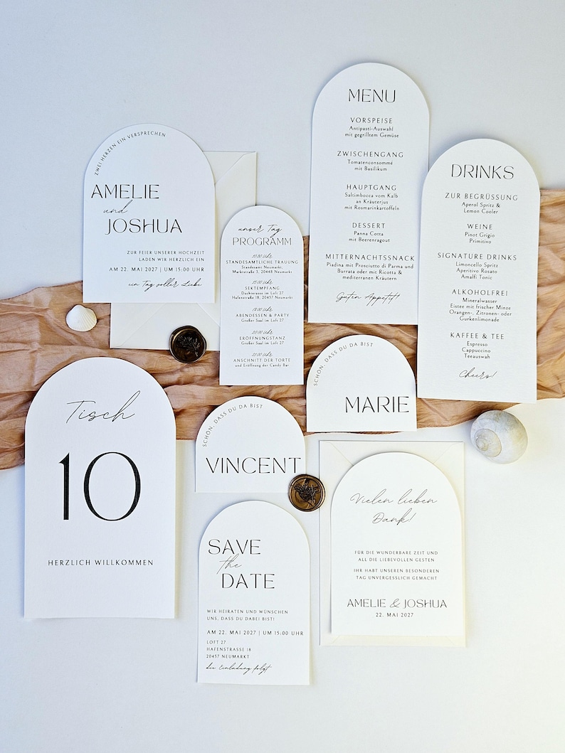 May include: A collection of white wedding stationery with black text, including invitations, menus, and table cards. The cards feature arched tops and elegant fonts. Decorative elements include wax seals and a seashell.
