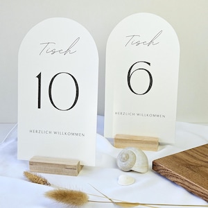 May include: Two arched table number cards with the word "Tisch" and numbers 10 and 6 in black script. The cards are on wooden stands, with the words "HERZLICH WILLKOMMEN" printed below the numbers. Decorative elements include a seashell and dried plants.