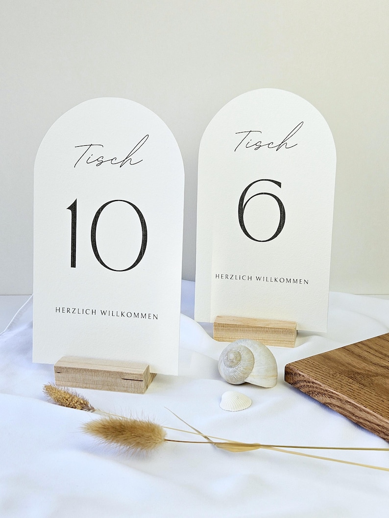 May include: Two arched table number cards with the word "Tisch" and numbers 10 and 6 in black script. The cards are on wooden stands, with the words "HERZLICH WILLKOMMEN" printed below the numbers. Decorative elements include a seashell and dried plants.