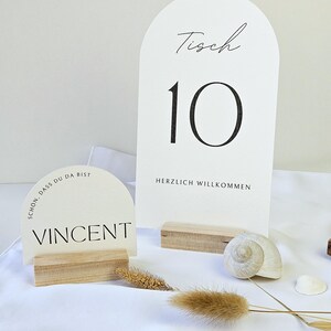 May include: White table number cards with black text, including the number 10 and the word "Tisch". A smaller card displays the name "Vincent". The cards are set on wooden bases, with decorative seashells and dried plants.