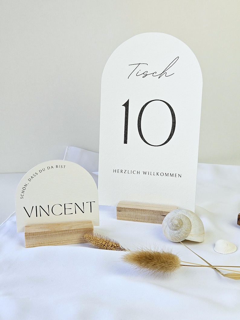 May include: White table number cards with black text, including the number 10 and the word "Tisch". A smaller card displays the name "Vincent". The cards are set on wooden bases, with decorative seashells and dried plants.