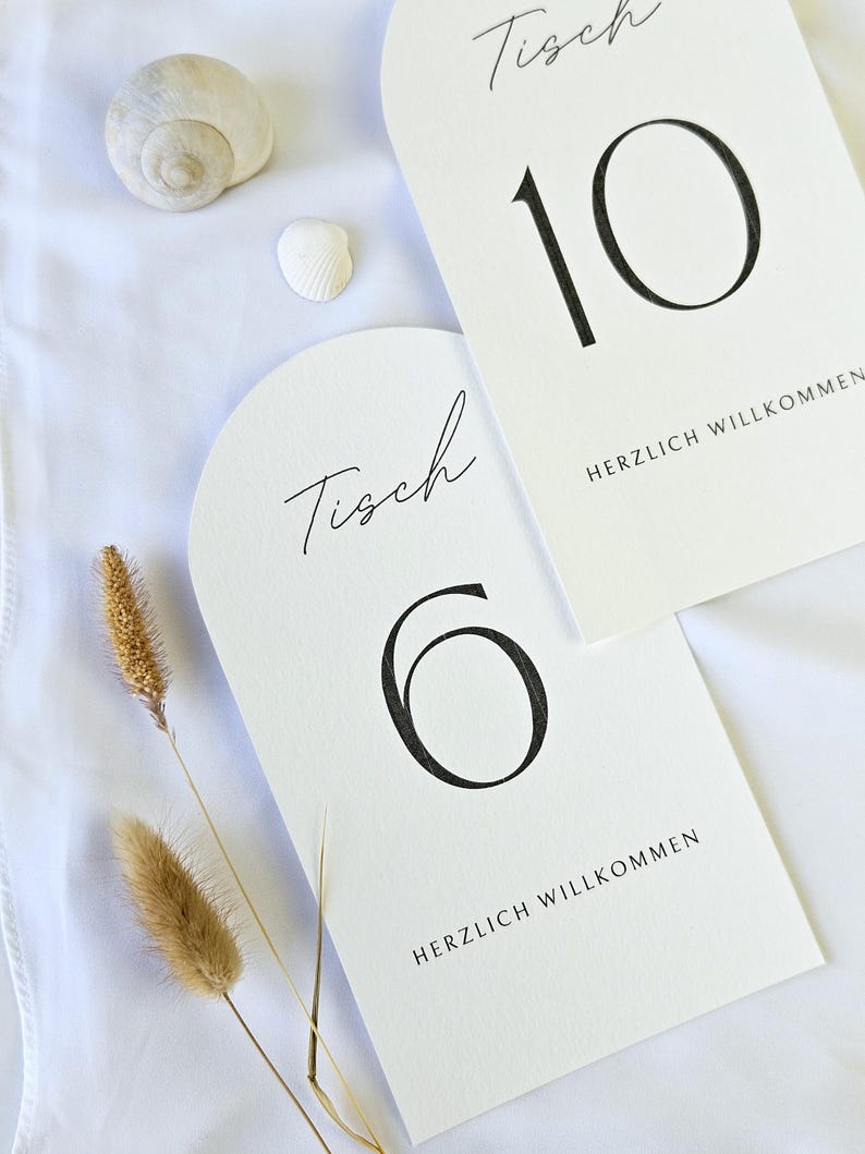 May include: Two white arched table number cards with black numerals 6 and 10. The word "Tisch" is written in script above each number. The phrase "HERZLICH WILLKOMMEN" is printed below the numbers. Seashells and dried grass are also visible.