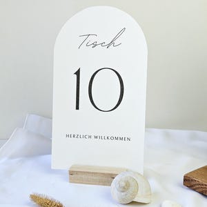 Può includere: Un segnaposto bianco con la parola "Tisch" e il numero "10" in corsivo nero. Il segnaposto è tenuto da un piccolo blocco di legno. Le parole "HERZLICH WILLKOMMEN" sono stampate in basso. Una conchiglia e piante secche in primo piano.