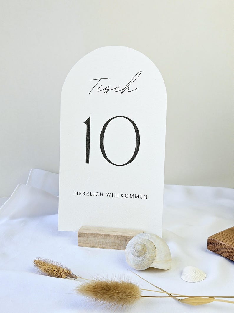 May include: A white table number card with the number 10 in a bold, black font. The word "Tisch" is written in cursive at the top, and "HERZLICH WILLKOMMEN" is at the bottom. The card is displayed on a wooden base, with a seashell and dried plants nearby.