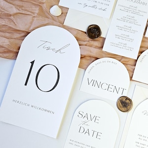May include: Elegant wedding stationery set featuring white cards with black calligraphy. Includes a table number, invitation, and program, accented with wax seals. The table number reads "Tisch 10" and "Herzlich Willkommen".