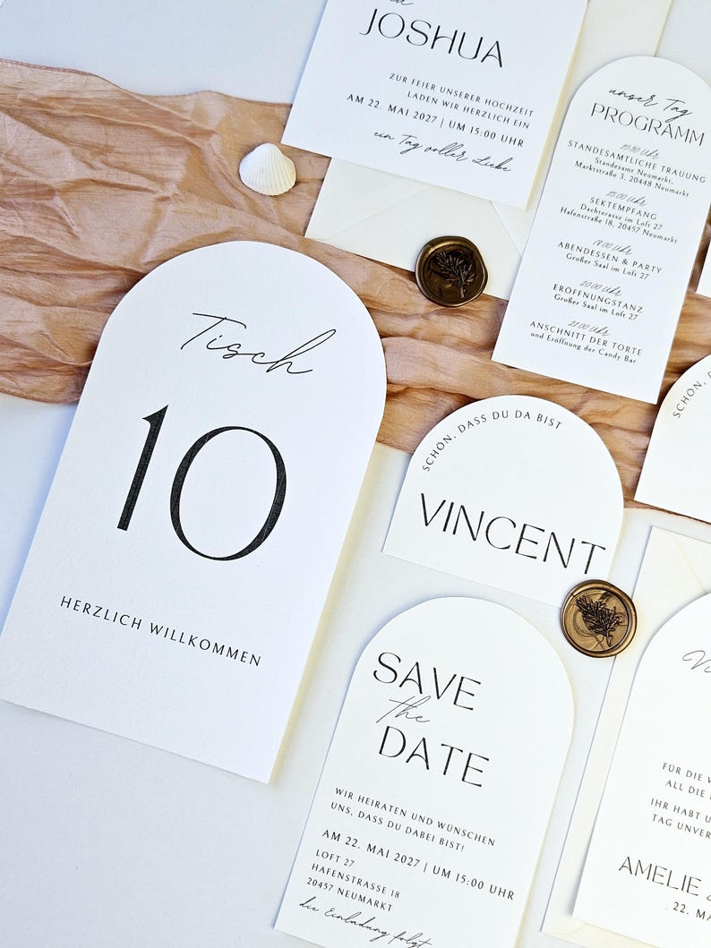 May include: Elegant wedding stationery set featuring white cards with black calligraphy. Includes a table number, invitation, and program, accented with wax seals. The table number reads "Tisch 10" and "Herzlich Willkommen".