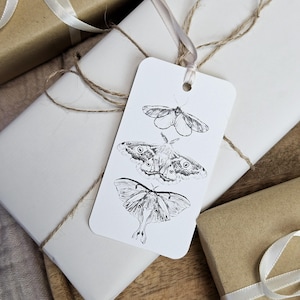 May include: White gift tag with three detailed moth illustrations in black ink. The tag is attached to a wrapped gift with twine and a cream-colored ribbon. Other wrapped gifts are visible in the background.