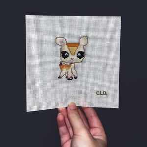 May include: A cross-stitch design of a deer-like creature with large black eyes, tan and brown fur, and a small purple bow. The design is on a white canvas with a grid pattern. The initials "CLD" are in the bottom right corner.