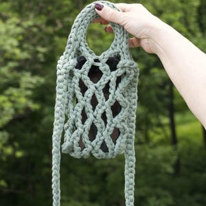 May include: A sage green crochet phone bag featuring an open diamond-patterned mesh design made from thick cord. The bag includes a top handle and a long braided shoulder strap, with a black rectangular smartphone visible inside the mesh structure.