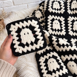 Puede incluir: Un cuadrado de ganchillo tipo granny square que presenta un motivo central de fantasma blanco sobre un fondo negro, rodeado por un borde blanco. El patrón se repite en una manta más grande, con cada cuadrado mostrando el mismo diseño.