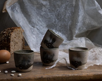 May include: Four handmade ceramic cups with a textured, speckled finish. The cups are a muted gray with dark brown accents near the base. The cups are arranged on a rustic wooden surface, with bread, an egg, and beans in the background.