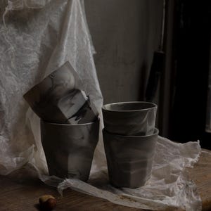 May include: Four gray and white marbled ceramic cups are stacked on a wooden surface. The cups have a geometric design and a smooth finish. Two Brazil nuts are on the wooden surface.