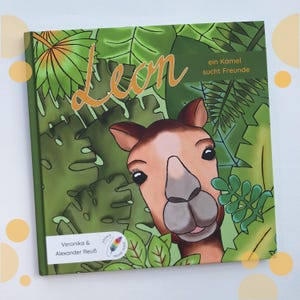 Könnte beinhalten: Ein Kinderbuch mit dem Titel "Leon" und einer Kamelillustration, die durch grünes Laub späht. Das Cover zeigt den Titel in orangefarbener Schrift, mit zusätzlichem Text auf Deutsch. Das Buch ist von Veronika & Alexander Reuß.