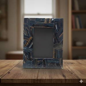 May include: A rectangular photo frame with a dark blue and gold stone mosaic design. The frame has a black interior and is displayed on a wooden table. The background includes a window and a bookshelf.