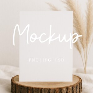 Könnte beinhalten: Eine weiße rechteckige Karte mit dem Wort "Mockup" in handschriftlichem Stil und dem Text "PNG | JPG | PSD". Die Karte liegt auf einer Holzscheibe, mit getrockneten Pflanzen im Hintergrund. Das Mockup Studio-Logo befindet sich unten.