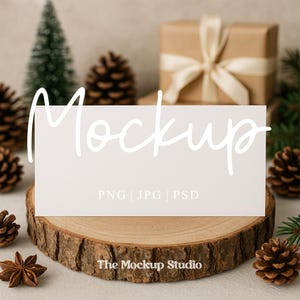 May include: A white rectangular card with the word "Mockup" in cursive script, resting on a wooden slice. The card displays "PNG | JPG | PSD" in a smaller font. Pine cones, a small Christmas tree, and a wrapped gift with a ribbon are in the background.
