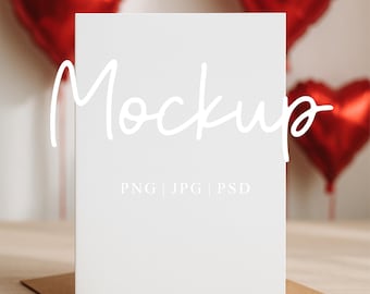 Puede incluir: Una tarjeta blanca con la palabra "Mockup" en una fuente de escritura, apoyada en un sobre marrón. Globos rojos en forma de corazón están en el fondo. El texto "PNG | JPG | PSD" está debajo de la palabra "Mockup". The Mockup Studio UK está en la parte inferior.