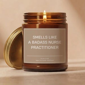May include: A lit brown glass jar candle with a gold lid. The candle has a white label with the text "SMELLS LIKE A BADASS NURSE PRACTITIONER". The candle is soy wax, vegan, eco-friendly, and hand-poured.