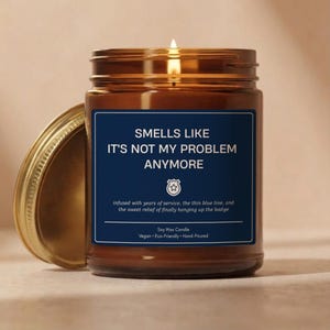 May include: A lit soy wax candle in a brown glass jar with a gold lid. The candle has a navy blue label with white text that reads "Smells Like It's Not My Problem Anymore". The candle is vegan, eco-friendly, and hand-poured.