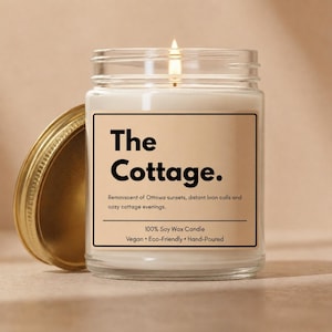 May include: A clear glass jar candle with a lit wick and a gold-colored metal lid placed to the side. The label features the text The Cottage. Reminiscent of Ottawa sunsets, distant loon calls and cozy cottage evenings. 100% Soy Wax Candle Vegan Eco-Friendly Hand-Poured.