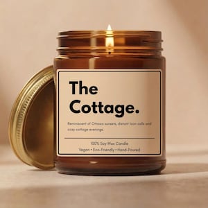 May include: A brown glass jar candle with a lit wick and a gold metal lid. The cream-colored label features the text The Cottage. Reminiscent of Ottawa sunsets, distant loon calls and cozy cottage evenings. 100% Soy Wax Candle Vegan Eco-Friendly Hand-Poured.