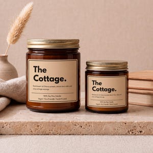May include: Two amber glass jars with gold lids, each featuring a beige label with the text The Cottage. The labels include the description Reminiscent of Ottawa sunsets, distant loon calls and cozy cottage evenings, 100% Soy Wax Candle, Vegan, Eco-Friendly, Hand-Poured.