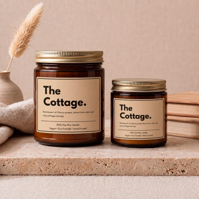 May include: Two amber glass jars with gold lids, each featuring a beige label with the text The Cottage. The labels include the description Reminiscent of Ottawa sunsets, distant loon calls and cozy cottage evenings, 100% Soy Wax Candle, Vegan, Eco-Friendly, Hand-Poured.