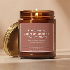 May include: A lit soy wax candle in a brown glass jar with a gold lid. The label reads "The Calming Scent of Escaping The Sh*t Show." The candle is vegan, eco-friendly, and hand-poured.