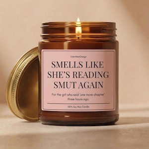May include: A brown glass jar candle with a lit wick and a gold lid placed to the side. The label is light pink with black text that reads: EdenNestDesign, SMELLS LIKE SHE'S READING SMUT AGAIN, For the girl who said 'one more chapter' three hours ago, 100% Soy Wax Candle.